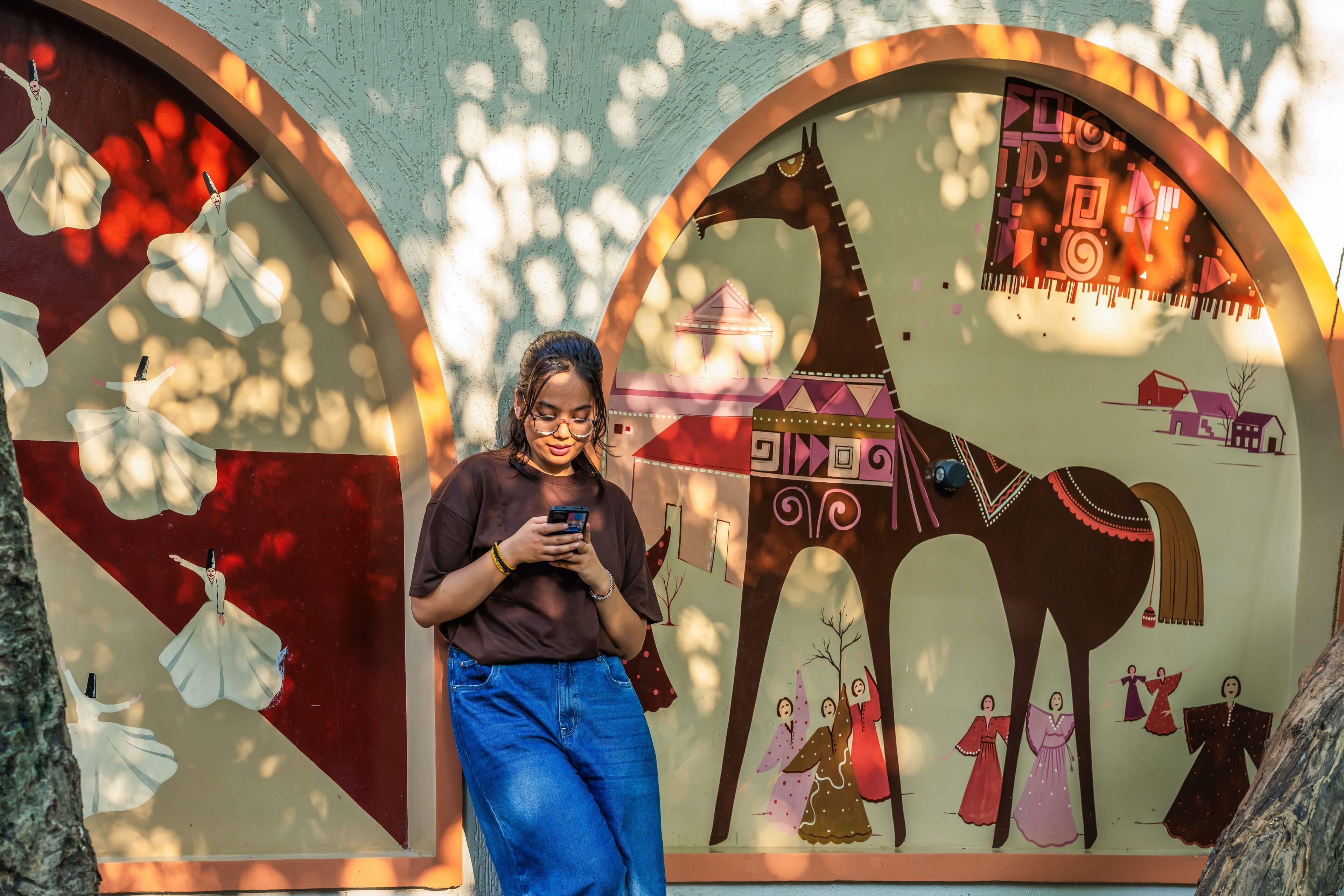 Una persona frente a un muro con murales coloridos, incluyendo un caballo y personas con vestimenta tradicional.