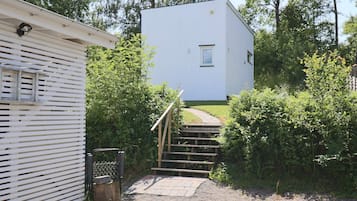 Un edificio blanco con una ventana pequeña y una puerta, rodeado de abundante vegetación y un sendero de grava que conduce a una escalera de madera.
