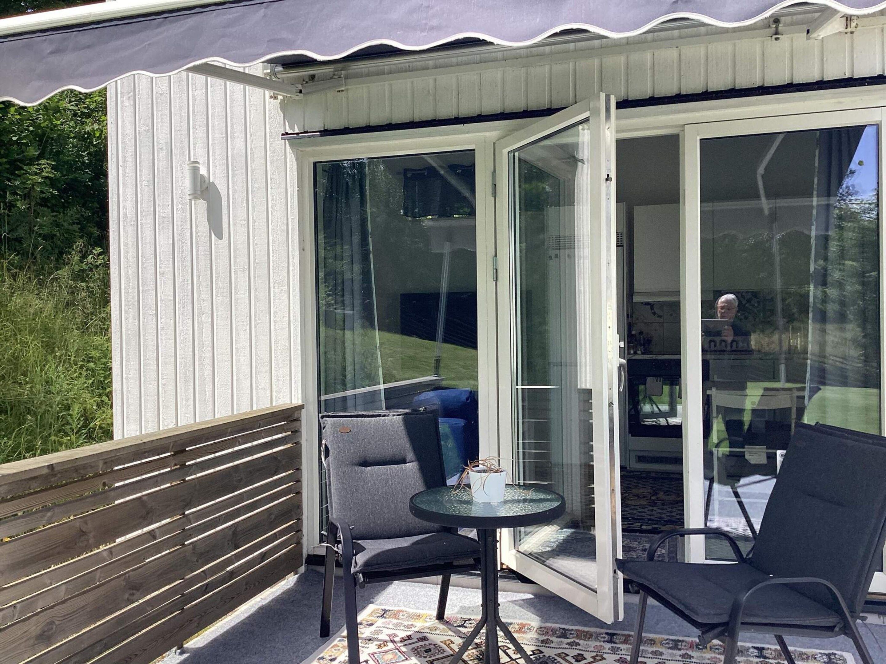 Un espacio de estar al aire libre con techo, que incluye una mesa y dos sillas, una cerca de madera y un portón de vidrio que da a un espacio interior.