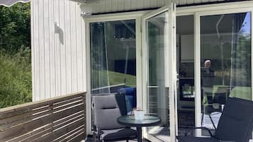Un espacio de estar al aire libre con techo, que incluye una mesa y dos sillas, una cerca de madera y un portón de vidrio que da a un espacio interior.