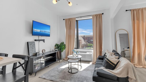 A modern living room with a flat-screen TV, a dining area, a sofa, and a large window with curtains. - Modern luxe apt Airport+ NYC (Elizabeth)