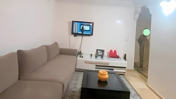 Una sala moderna con un sofá beige, una mesa de centro negra y una lámpara de araña decorativa. Un televisor de pantalla plana está montado en la pared, y se ve un aparador blanco con zapatos rojos.