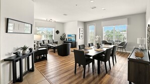 A spacious living room with a large dining table, black chairs, and a black sofa. There is a TV mounted on the wall, a fireplace, and a large window with a view of the outdoors.