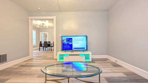 Un salon moderne avec une table basse en verre, un téléviseur à écran plat posé sur un meuble, et un coin repas à l’arrière-plan.