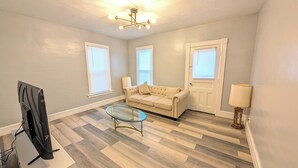 Un salon avec un canapé beige, une table basse en verre et un téléviseur posé sur un meuble. Il y a trois fenêtres équipées de stores, une porte et une lampe sur une table d’appoint.