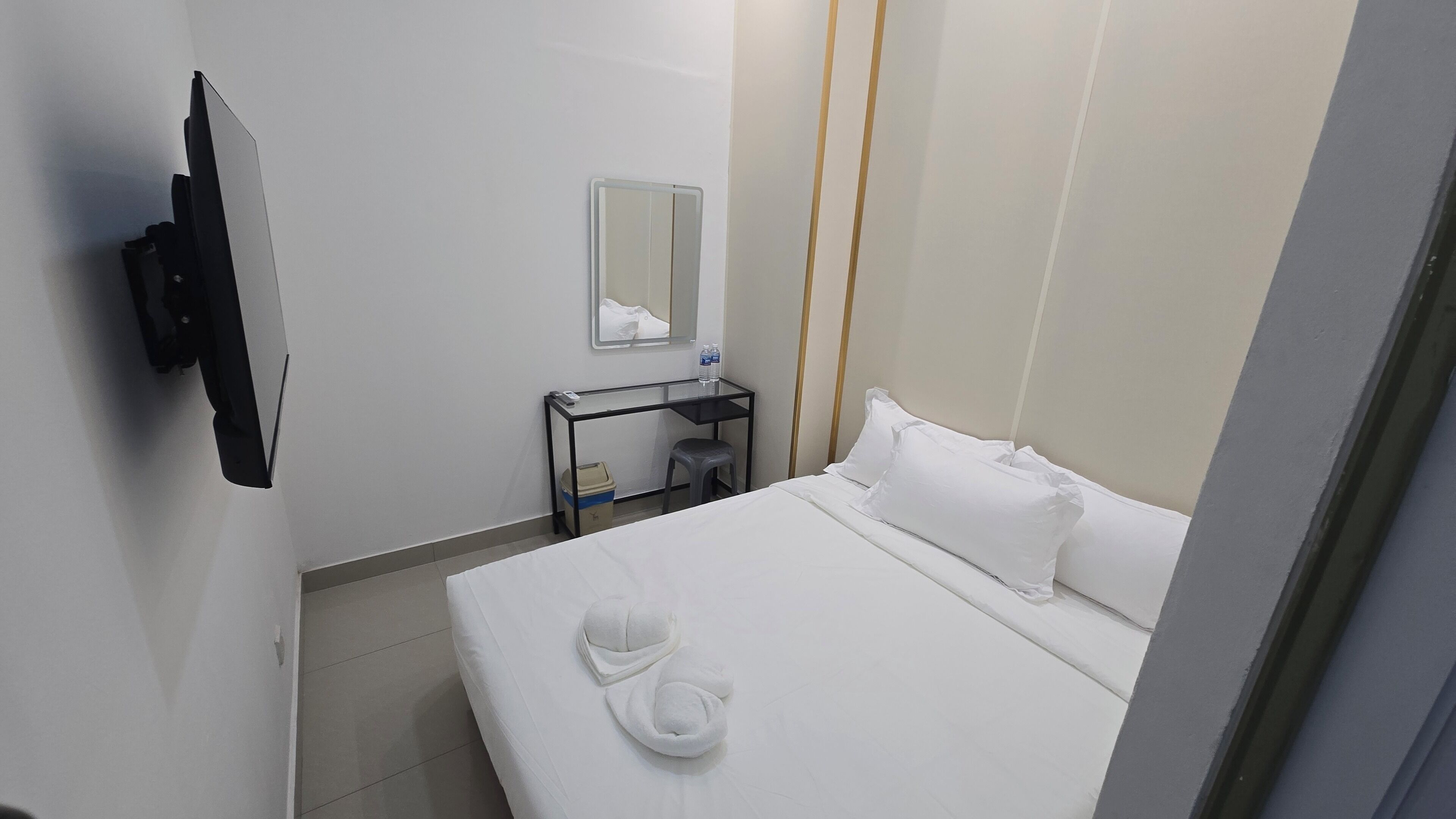A hotel room with a bed, a desk with a mirror, a small stool, and a TV mounted on the wall.