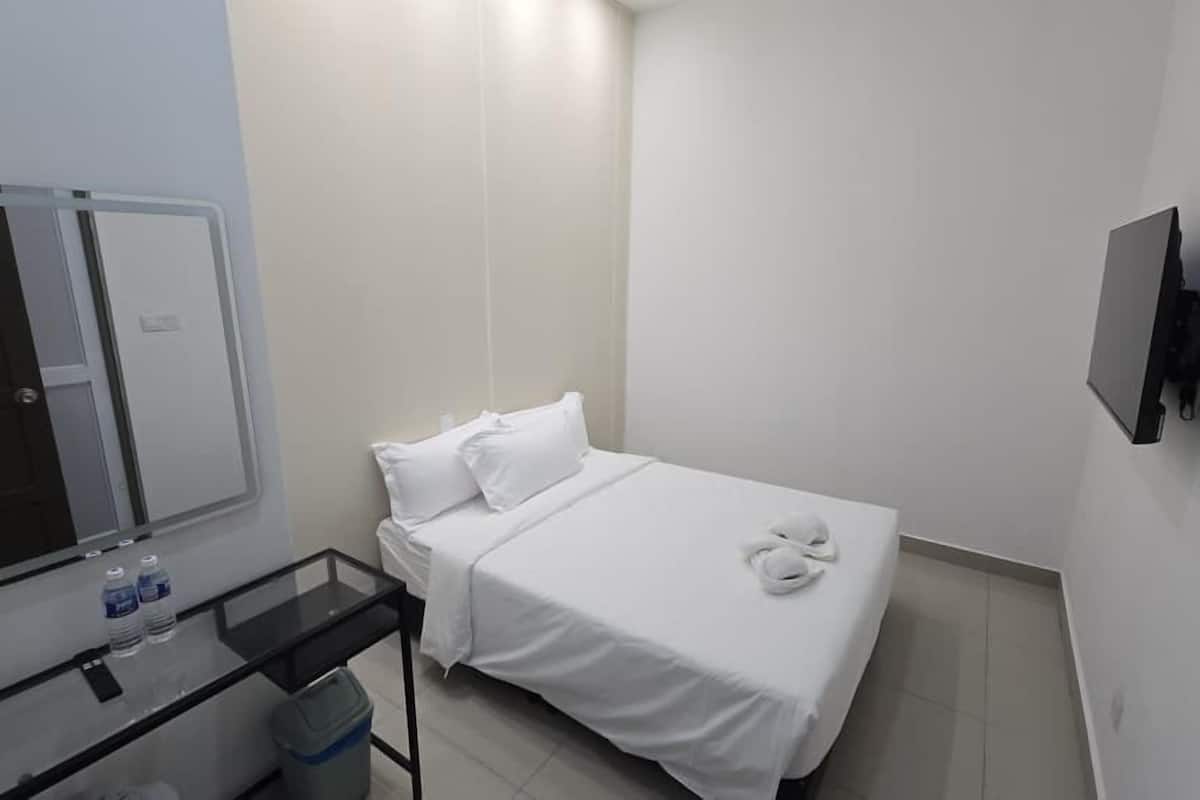 A small, clean room with a bed, a mirror, a TV, and a small table with a trash bin.