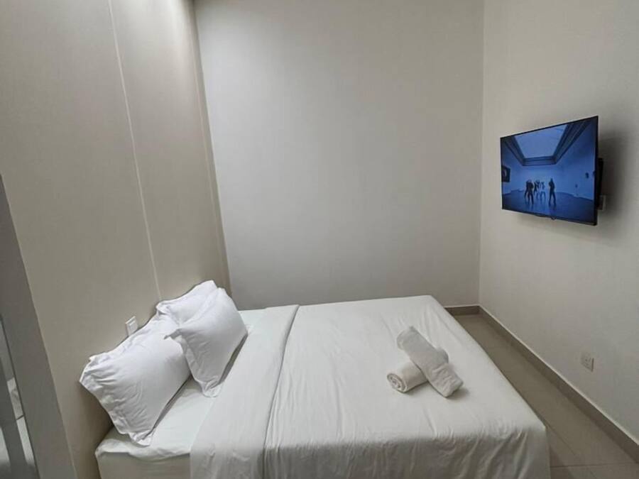 Quarto de hotel pequeno e moderno, com uma cama de solteiro, televisão de tela plana na parede e uma mesinha.
