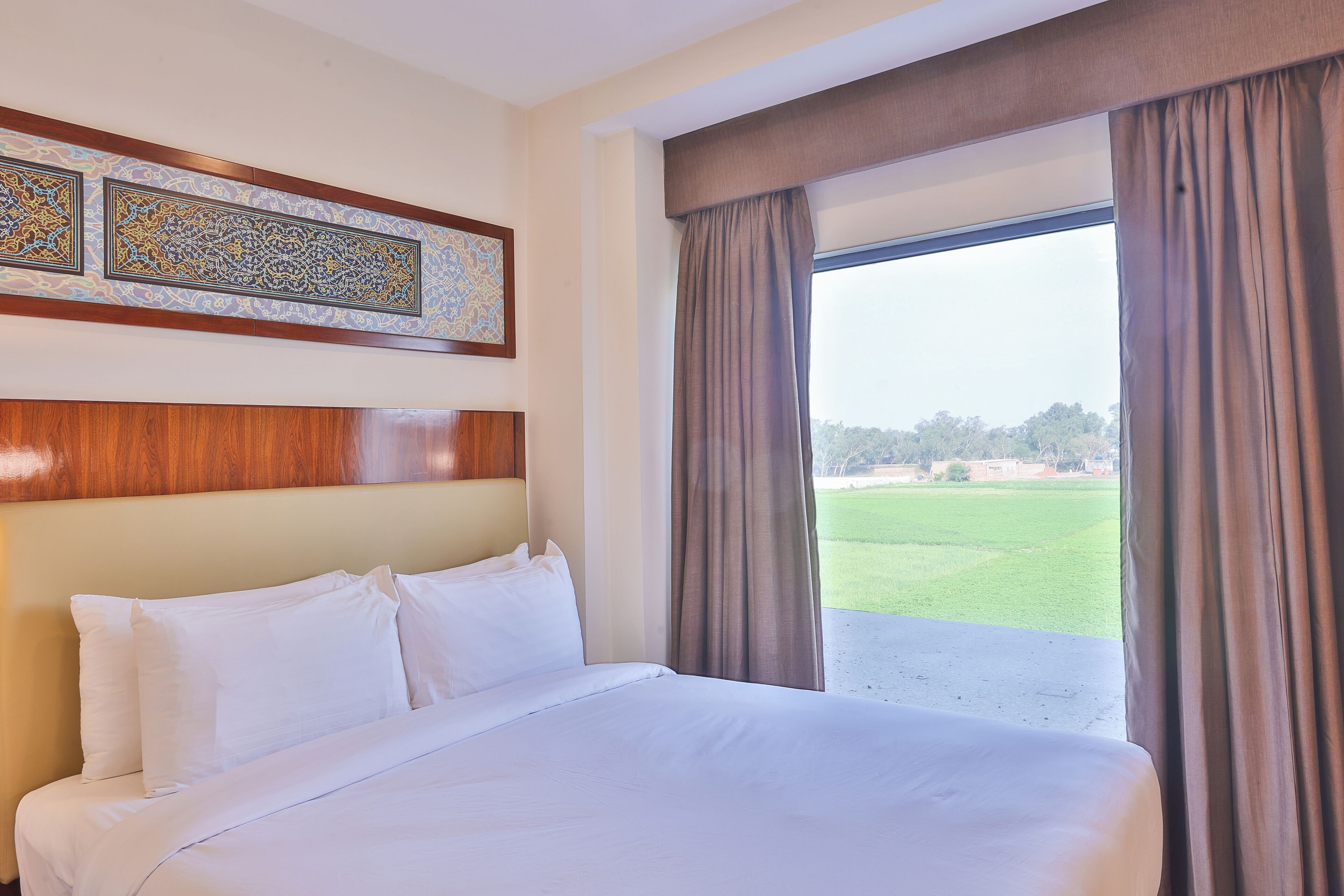A hotel room with a large bed, a view of a green field, and a decorative wall panel.