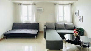 A modern living room with a sofa, ottoman, and a small table. There are two windows with curtains, an air conditioning unit on the wall, and a decorative plant on the table. - Spacious & Relaxing Haven (Iloilo)