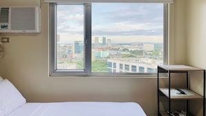A hotel room with a bed, a window with a view of the city, and a small shelf with a remote control. - The Perfect Central Getaway (Iloilo)