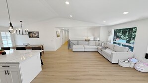 Een moderne woonruimte met een keukeneiland, een grote witte bank en uitzicht op groen door een raam.