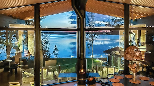 A modern interior with large windows overlooking a lake, a central fireplace, and a dining area with round tables. - Villa haave by Interhome (Kustavi)