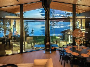A modern interior with large windows overlooking a lake, a central fireplace, and a dining area with round tables. - Villa haave by Interhome (Kustavi)