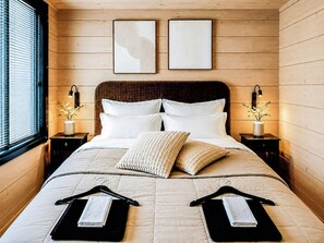 A bedroom with a large bed, two bedside tables, a window with blinds, and two wall-mounted picture frames.