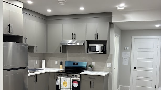 A modern kitchen with grey cabinets, a stainless steel refrigerator, a built-in microwave, and a stove. There is a grey sofa in the living area. - Cozy, clean and homely (Calgary)