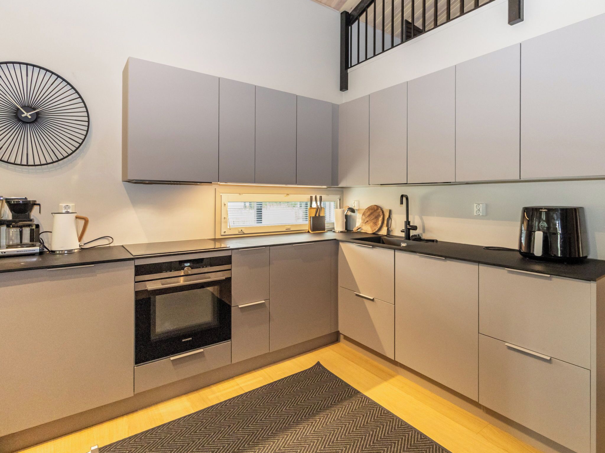 Een moderne keuken met grijze kastjes, een ingebouwde oven, een koffieapparaat en een grote wandklok.