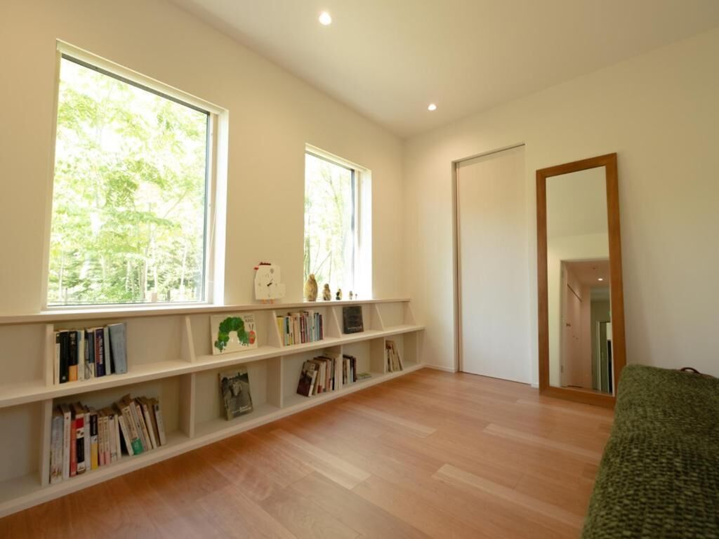 Una habitación amplia con piso de madera, una estantería llena de libros, dos ventanas grandes con vistas a árboles y un espejo en la pared.