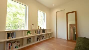 Una habitación amplia con piso de madera, una estantería llena de libros, dos ventanas grandes con vistas a árboles y un espejo en la pared.