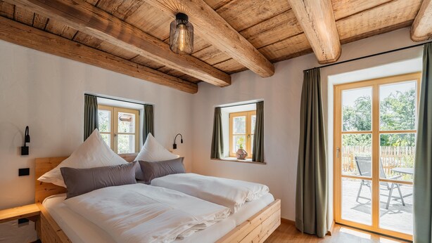 Un dormitorio con cama de madera, dos ventanas con cortinas, techo de madera y vistas al exterior.