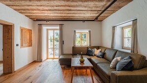 Una sala de estar acogedora con techo de madera, un sofá, una mesa de centro con frutero y dos ventanas con cortinas.
