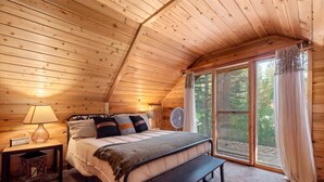 Un dormitorio acogedor con techo de madera, una cama con una banca, una mesita de noche con lámpara y un ventanal con cortinas.