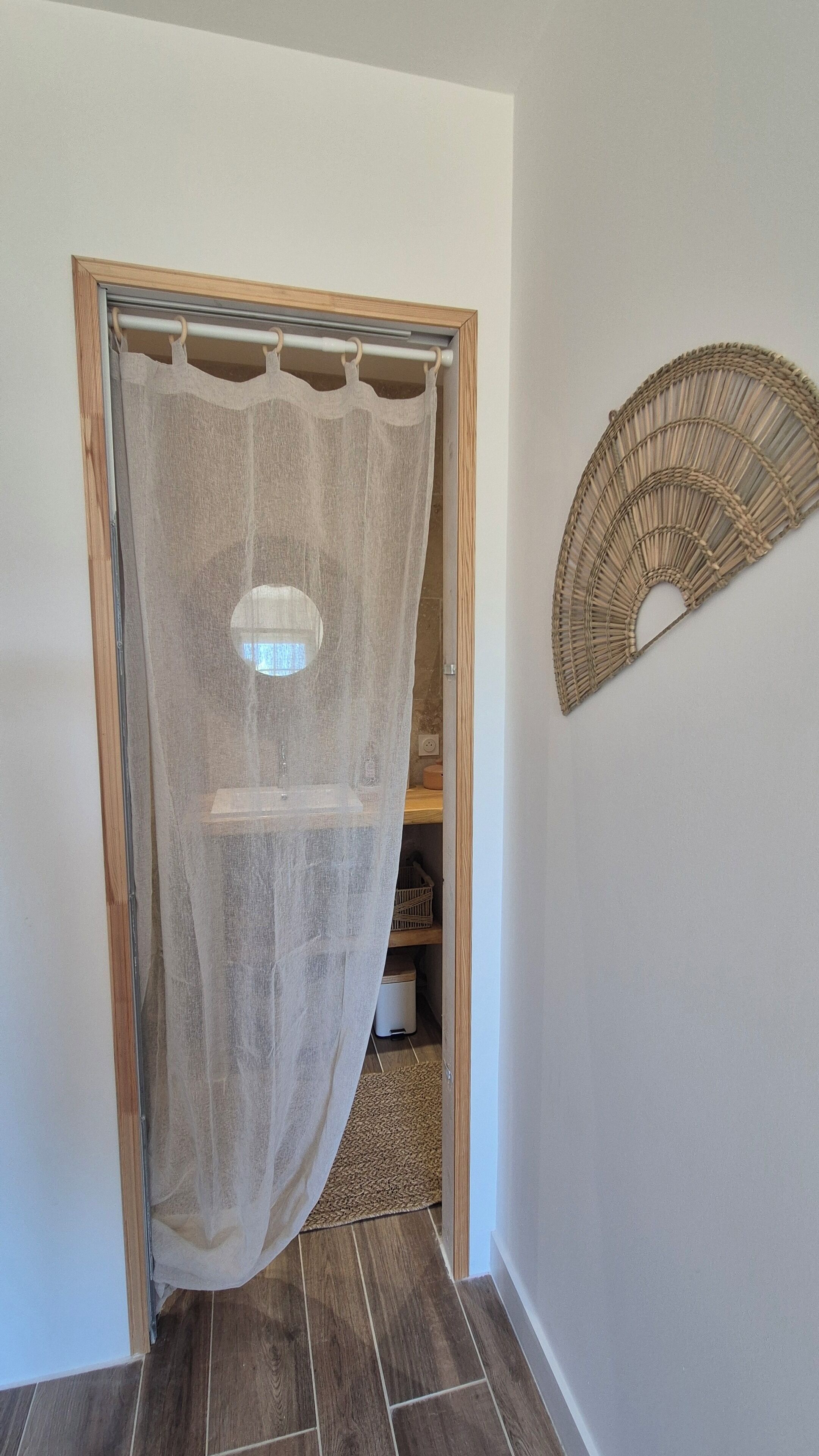 A hallway with a wooden door, a sheer curtain, a woven wall hanging, and a tiled floor.