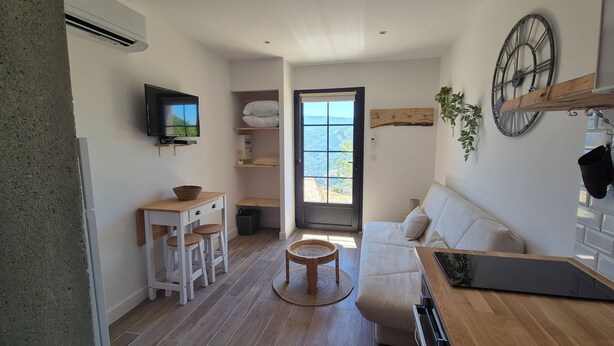 Un espace de vie compact comprenant un canapé, une petite table, un téléviseur et une vue sur l’extérieur, visible à travers une porte.