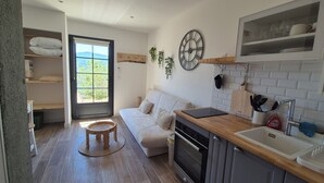 Une cuisine compacte dotée d’un four encastré, d’un évier et d’un plan de travail en bois. Une fenêtre offre une vue sur la verdure environnante.