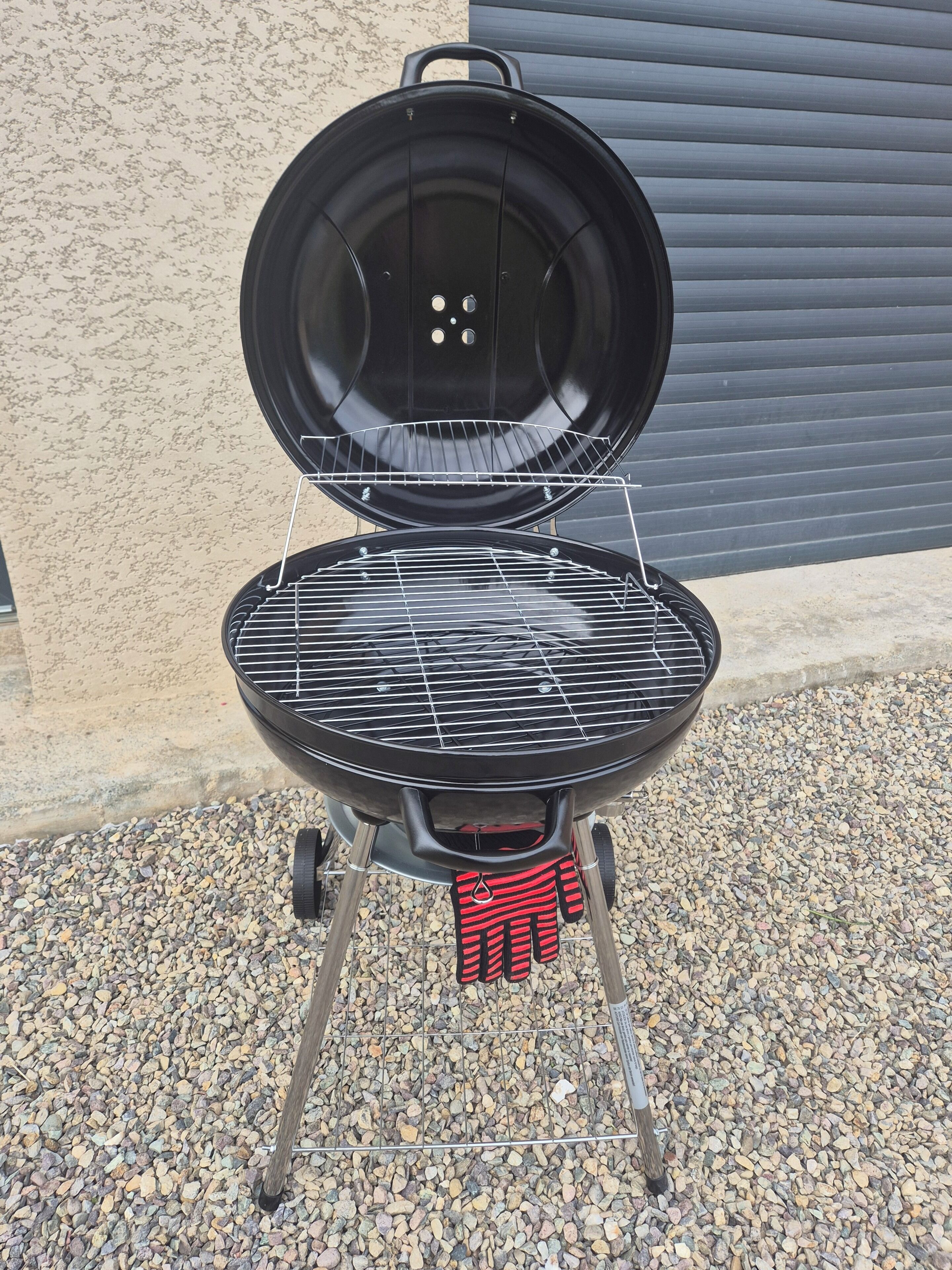 A portable grill with a lid open, placed on a gravel surface.