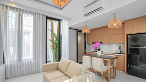 A modern living room with a sofa, dining table, and chairs. There is a kitchen area with wooden cabinets and a refrigerator.
