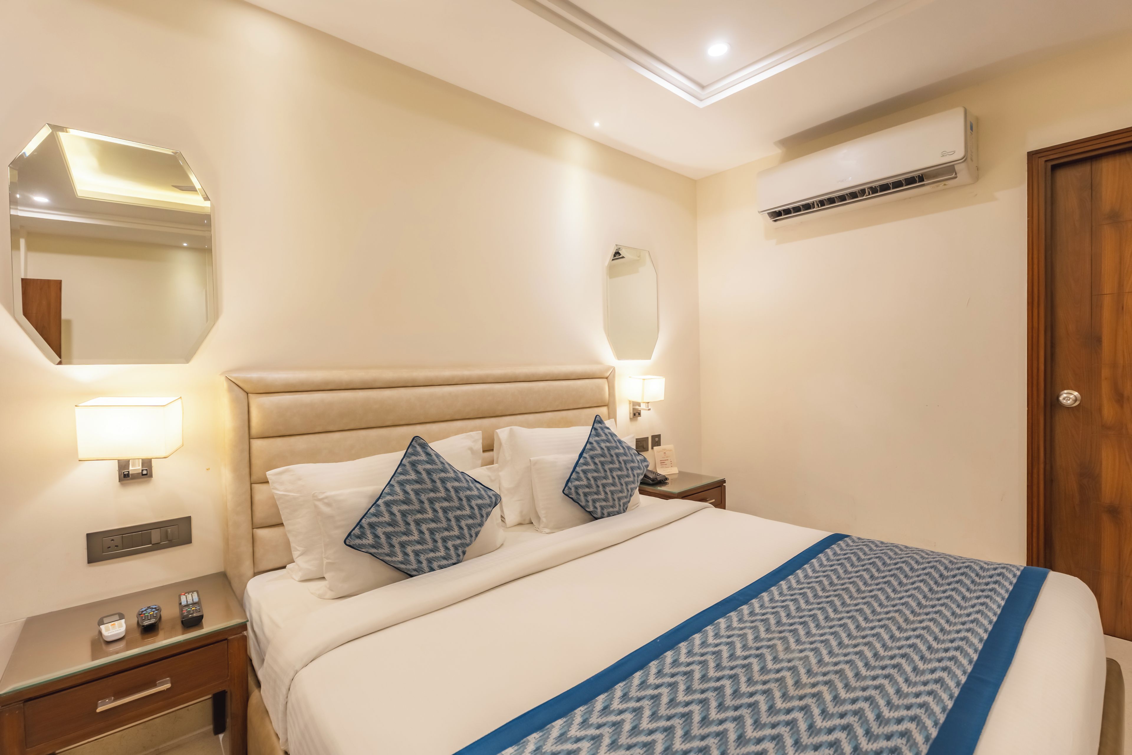 A hotel room with a bed, bedside tables, a nightstand, a mirror, an air conditioning unit, and a wooden door.