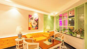 A room with a yellow sofa, two chairs, a table, a lamp, a painting, and a window with green shutters.