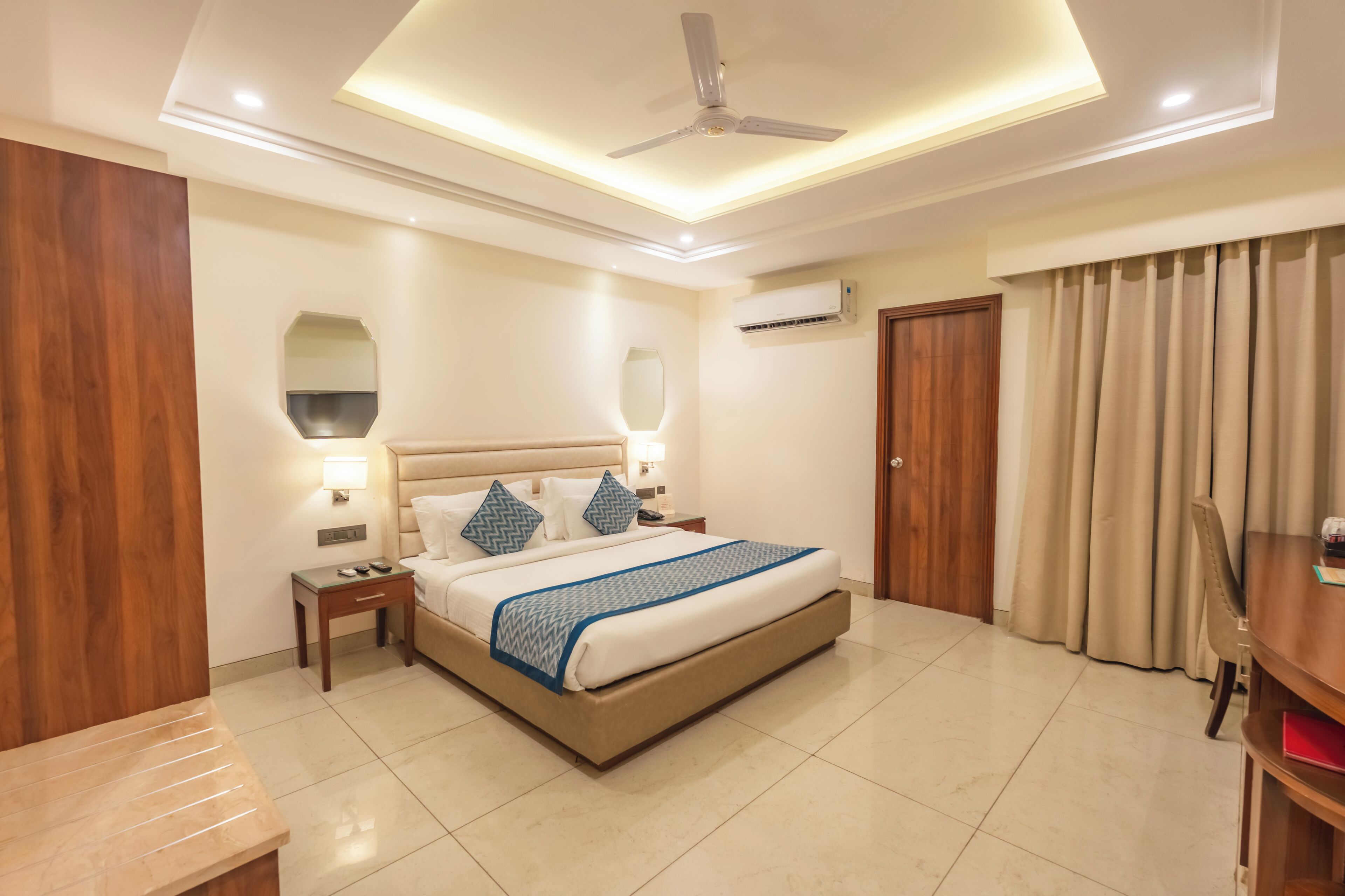 A hotel room with a bed, bedside tables, a ceiling fan, an air conditioning unit, and a door with curtains.