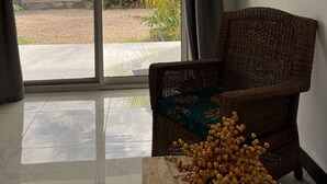 A room with a wicker chair, a wooden table with decorative items, and a sliding glass door leading to a balcony with a view of a garden and buildings.