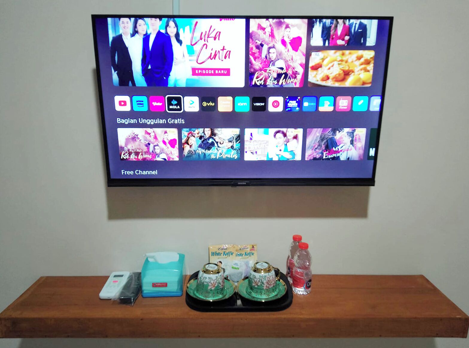 Ein Fernseher zeigt eine Streaming-Dienstanwendung mit verschiedenen Serien-Vorschaubildern und einem Hinweis auf einen 'kostenlosen Kanal'. Darunter ein Holztisch mit einem Smartphone, einer Taschentuchbox, einem kleinen dekorativen Gegenstand und einer Wasserflasche.