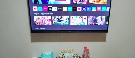 Ein Fernseher zeigt eine Streaming-Dienstanwendung mit verschiedenen Serien-Vorschaubildern und einem Hinweis auf einen 'kostenlosen Kanal'. Darunter ein Holztisch mit einem Smartphone, einer Taschentuchbox, einem kleinen dekorativen Gegenstand und einer Wasserflasche.