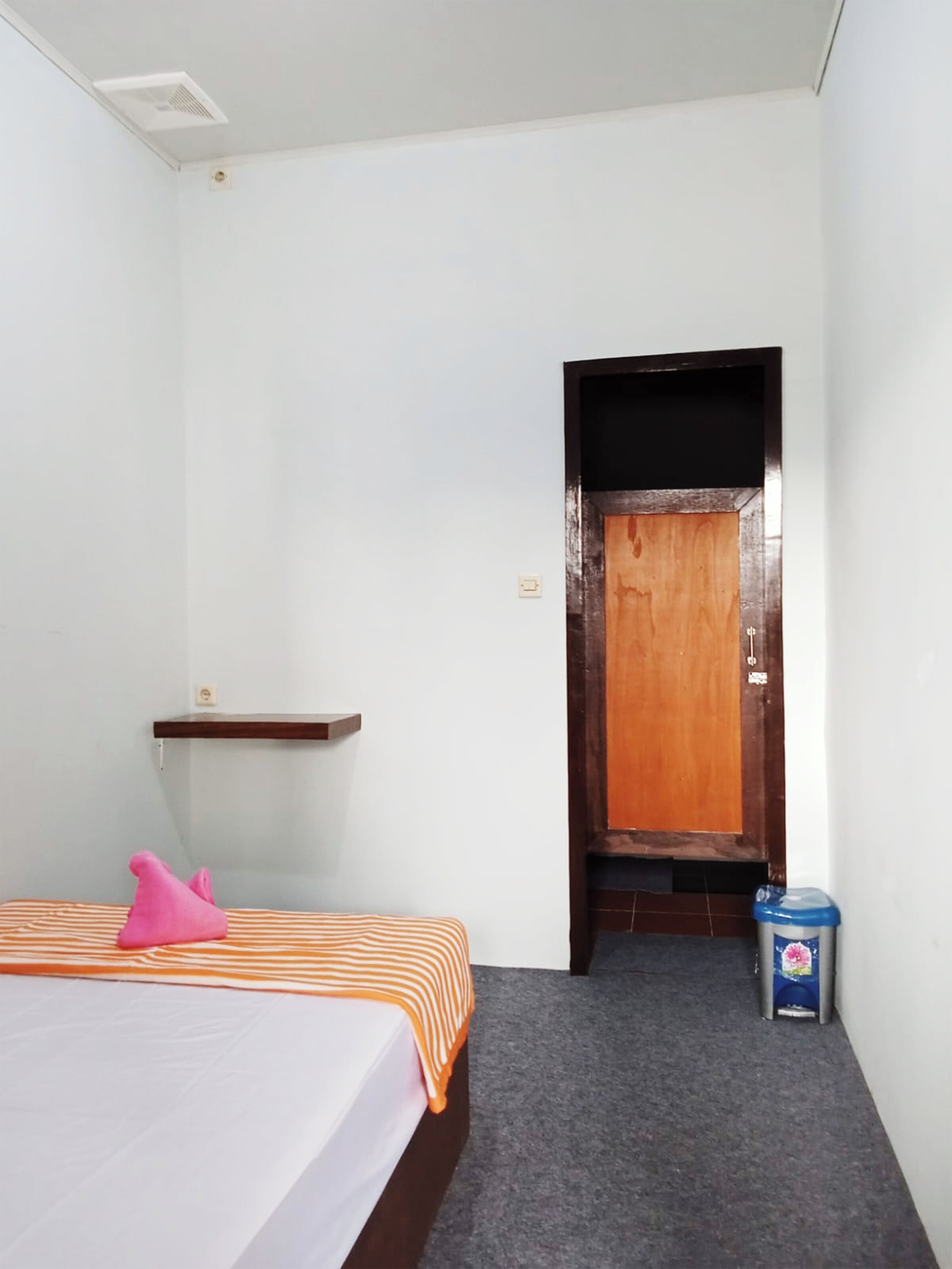 A small, clean room with a bed, a wooden door, a trash bin, and a small shelf.