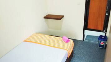 A small, single-bed room with a striped bedspread, a small wooden shelf, and a door leading to another room.