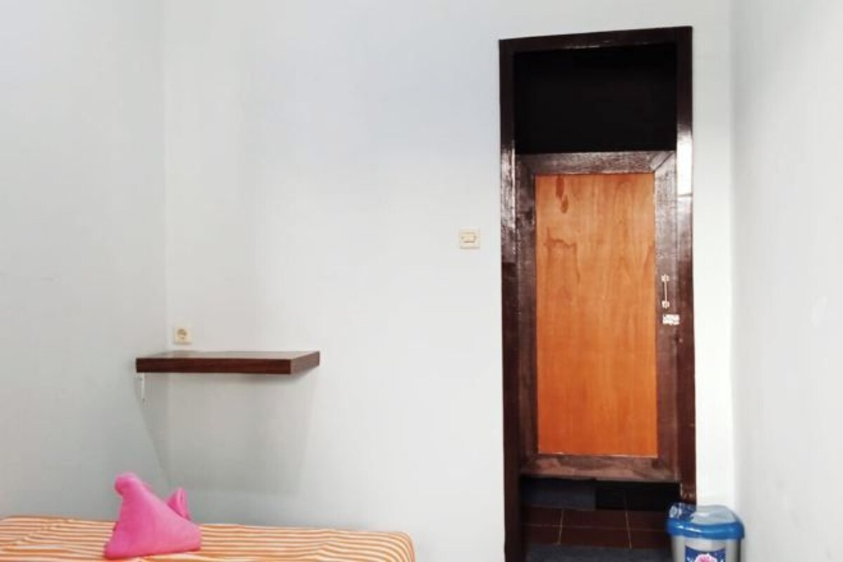 A room with a bed, a pink object on the bed, a wooden door, and a trash bin.