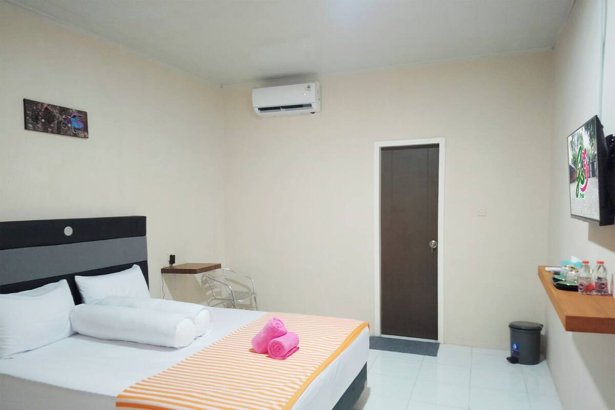 A hotel room with a bed, a small table, an air conditioning unit, a door, and a trash bin.