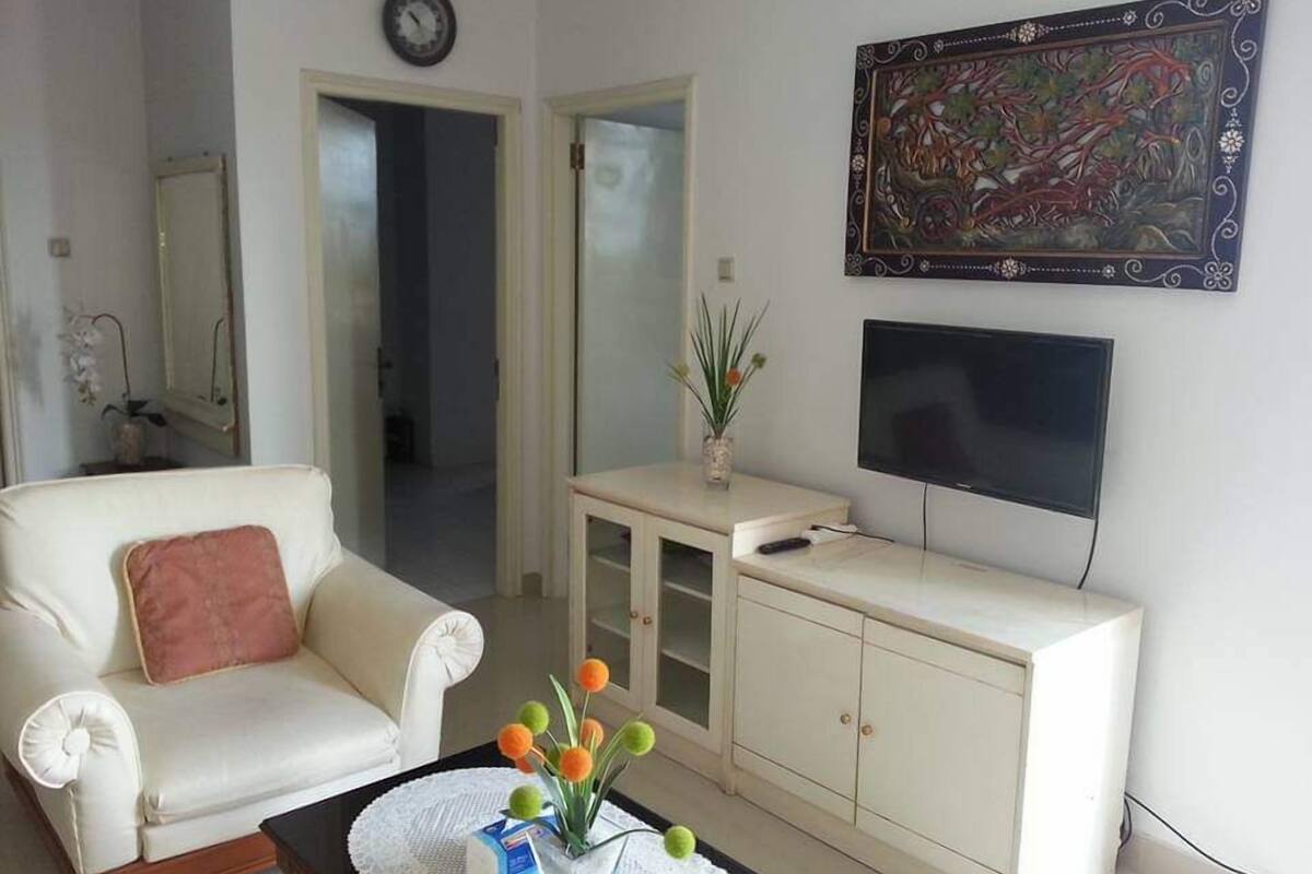Een woonkamer met een witte bank, een bijzettafeltje met een bloevas, een televisie op een standaard, en een kast.