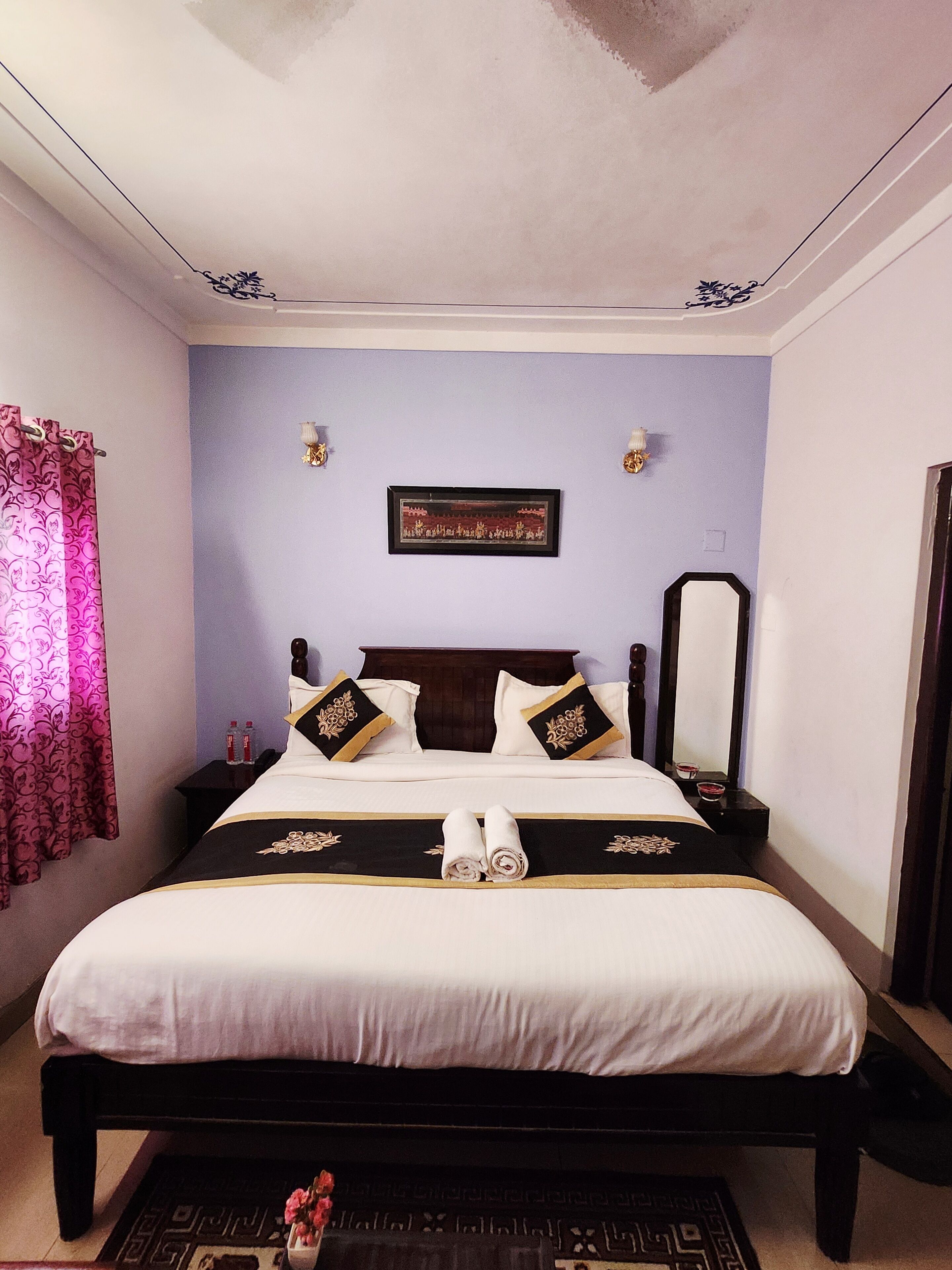 A neatly made bed with a black and gold bedspread, two pillows with similar designs, a framed picture on the wall, and a mirror on the wall.