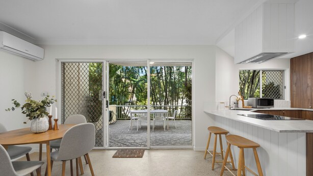 A modern kitchen with a white countertop, wooden stools, and a sliding glass door leading to an outdoor patio. - Burleigh Breeze 2 Bed Beach Walk (Burleigh Heads)