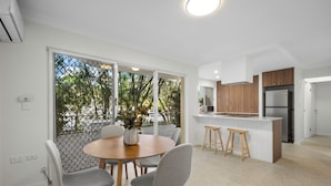 A modern kitchen with wooden cabinets, a stainless steel refrigerator, and a dining area with a round table and grey chairs. - Burleigh Breeze 2 Bed Beach Walk (Burleigh Heads)