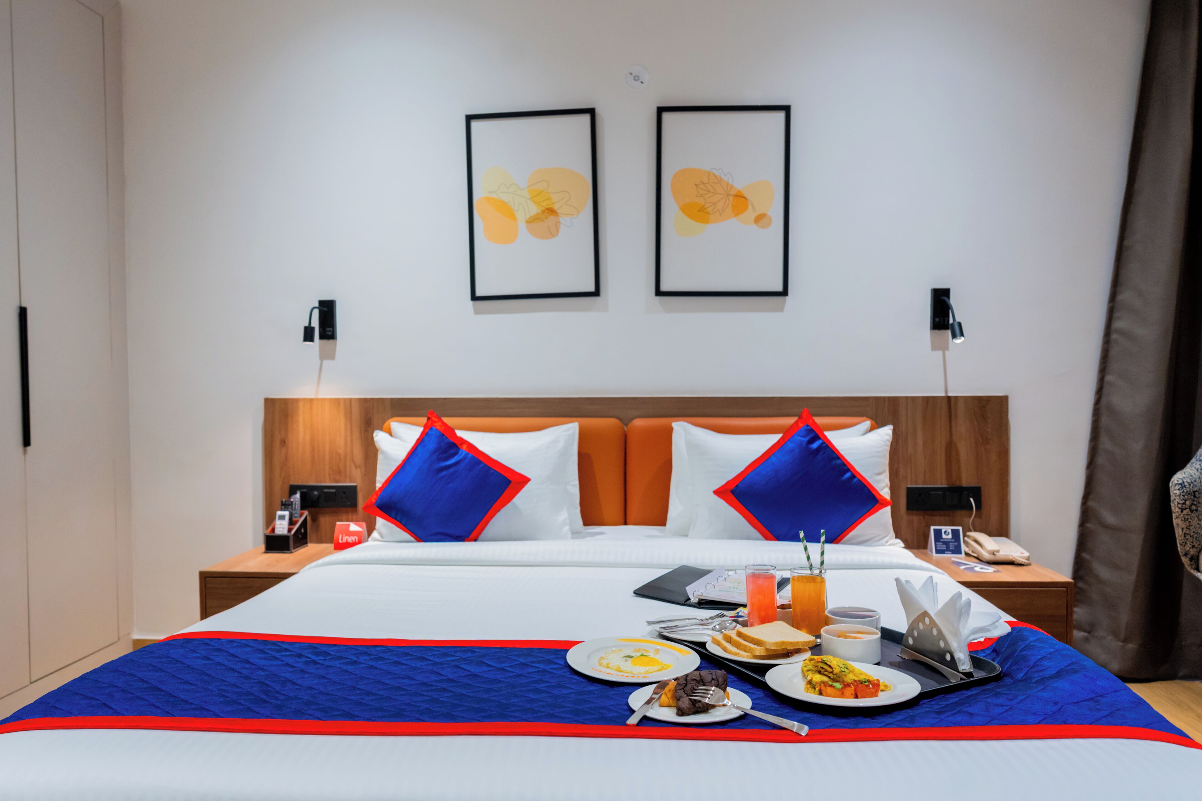 A hotel room with a bed, a bedside table with a lamp, a wall-mounted phone, and two framed pictures.