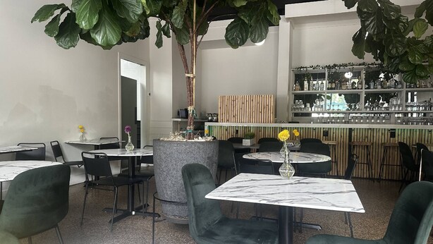 A restaurant with round tables, green upholstered chairs, and a bar area with shelves of bottles and glasses.