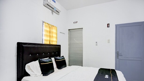 A hotel room with a bed, a window with curtains, an air conditioning unit, a door, and a wall-mounted light switch.