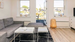 A modern living room with a grey sofa, a white coffee table, and a view of a canal and buildings through large windows.