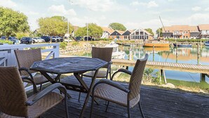 A wooden deck with wicker chairs and a table overlooking a marina with boats and buildings in the background.